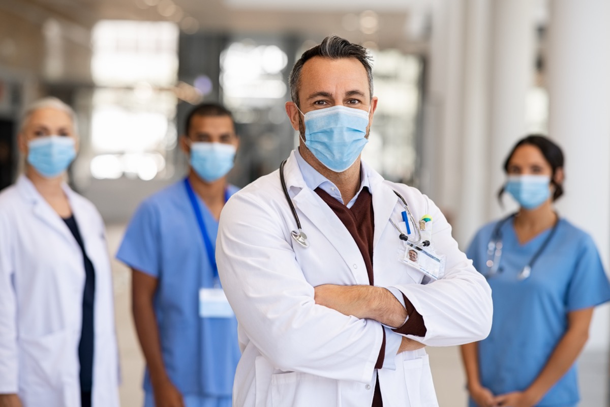 Mature doctor standing in corridor with medical team at hospital wearing surgical face mask due to covid. Smiling general practitioner with crossed arms looking at camera.