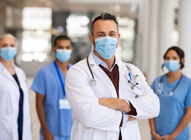 Mature doctor standing in corridor with medical team at hospital wearing surgical face mask due to covid. Smiling general practitioner with crossed arms looking at camera.