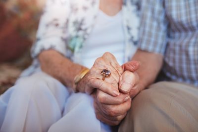 Cropped shot of elderly couple holding hands while sitting together at home. Focus on hands.