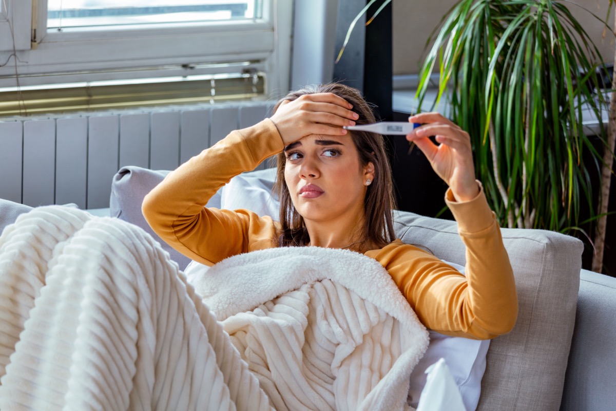 Woman being sick having flu lying on sofa looking at temperature on thermometer. Sick woman lying in bed with high fever.