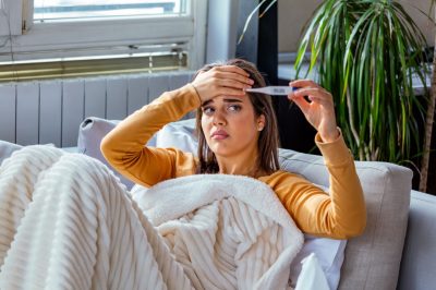 Woman being sick having flu lying on sofa looking at temperature on thermometer. Sick woman lying in bed with high fever.