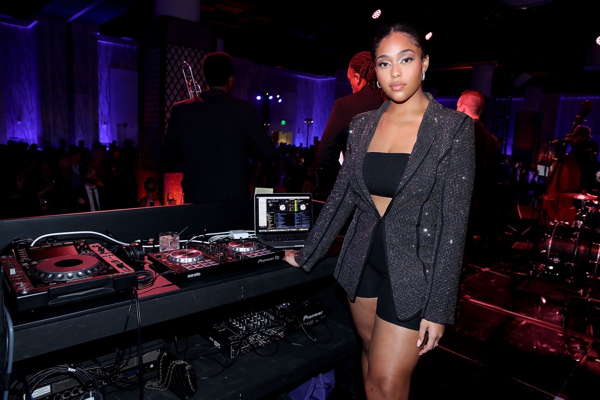 jordyn woods standing at dj booth in sparkly jacket, bandeau, and bike shorts