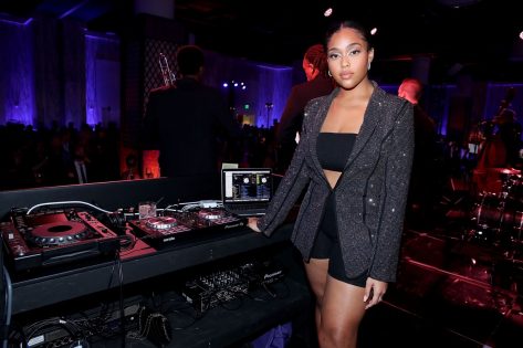 jordyn woods standing at dj booth in sparkly jacket, bandeau, and bike shorts
