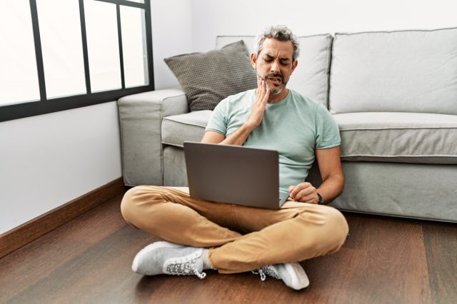 Middle age hispanic man using laptop sitting on the floor at the living room touching mouth with hand with painful expression.