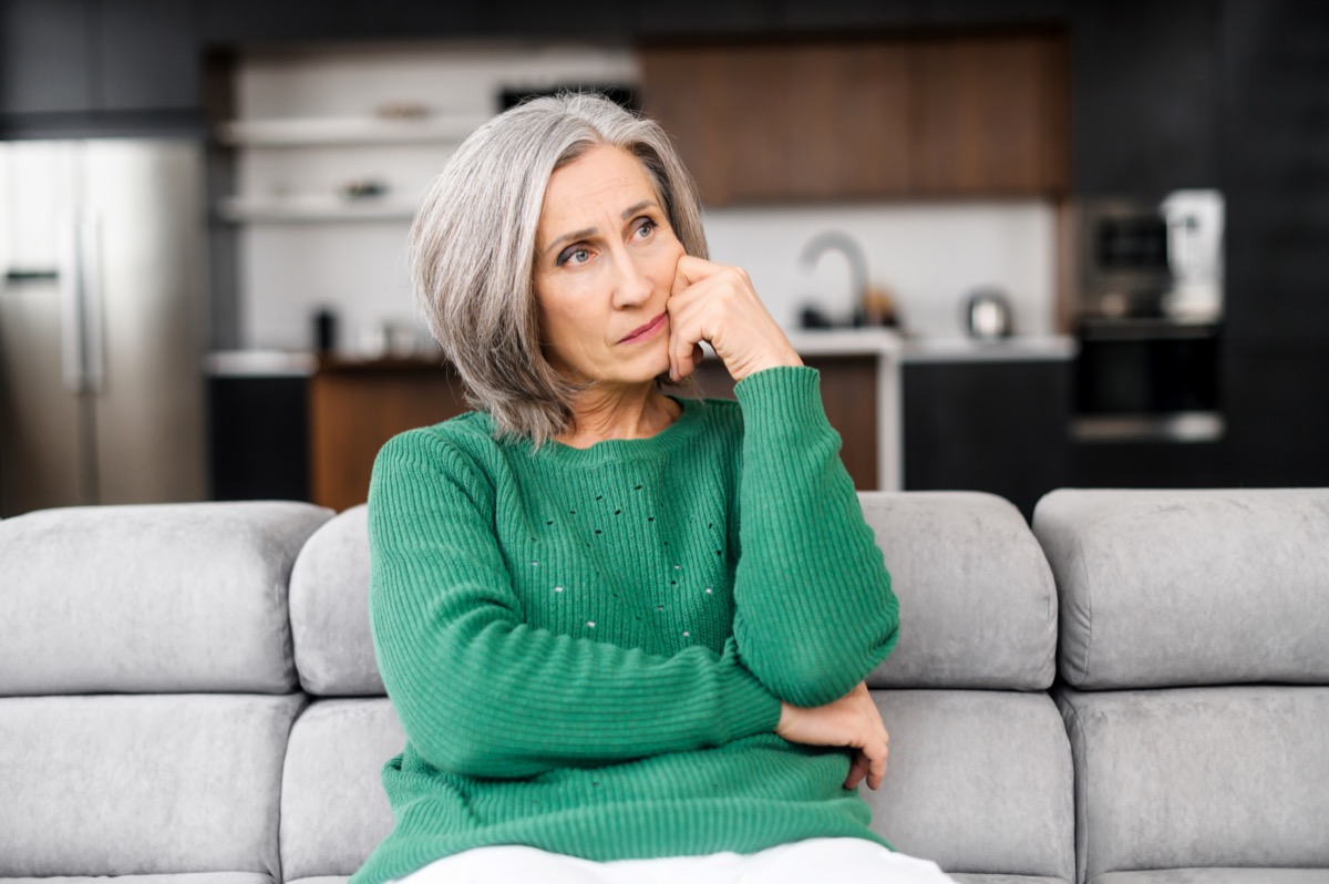 Mature woman with grey hair, thinking touching the face, looking aside, sitting on the pure sofa at apartment.