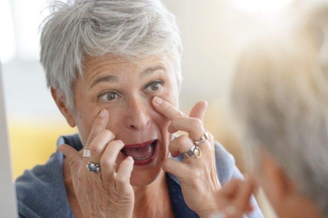 Mature white-haired woman checking eye wrinkles in front of mirror.