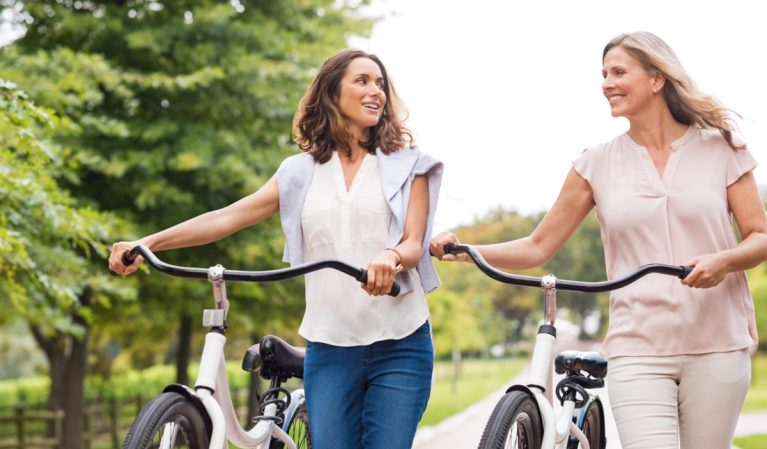 Two mature women in conversation while walking with bicycle at park. Happy beautiful senior women walking in the park with bicycles in a spring time. Friends holding bikes and talking to each other.