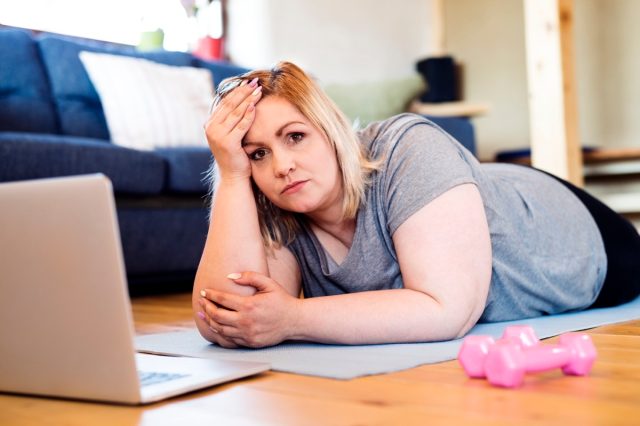 жена с наднормено тегло у дома лежи на пода, лаптоп пред нея, подготвена да тренира на постелка според видеото