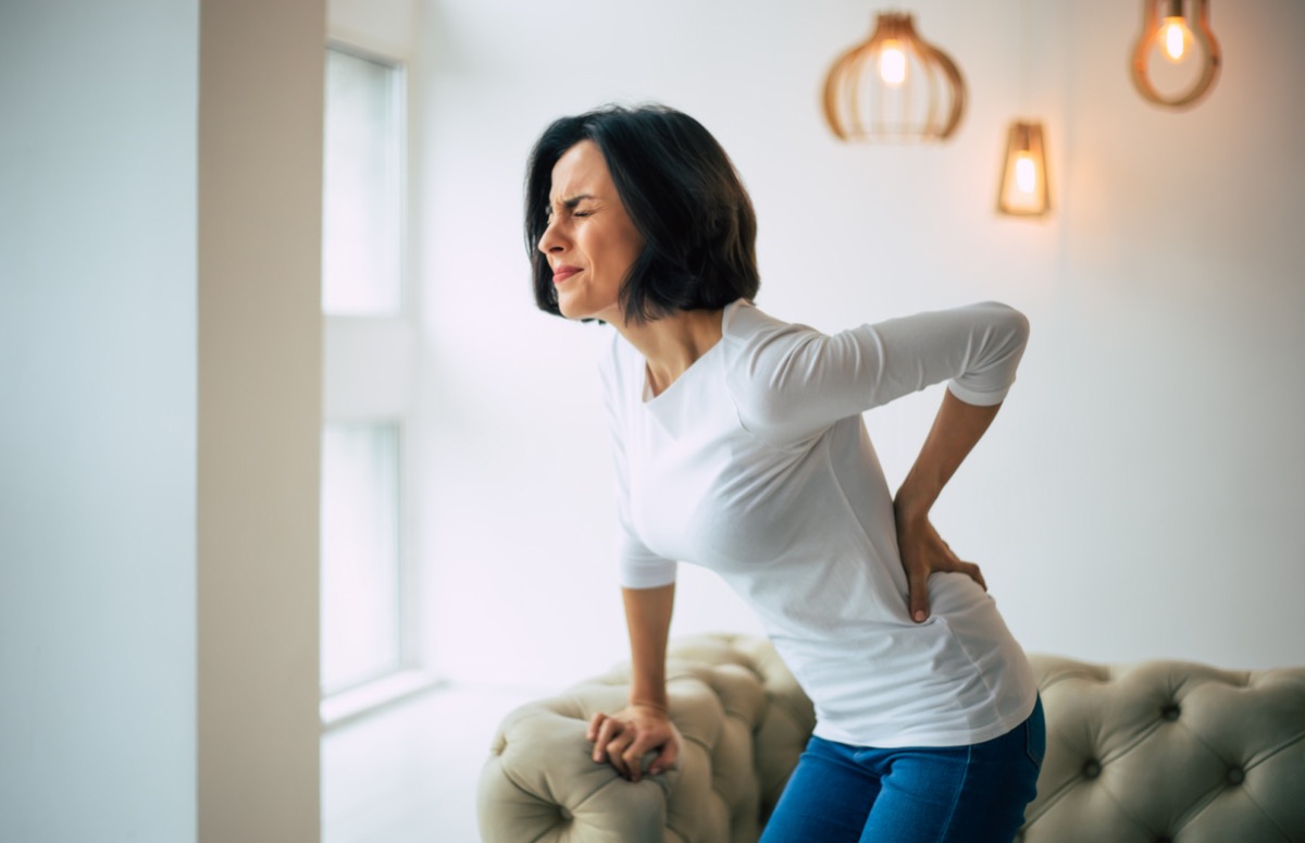 Woman is holding her lower back, while standing and suffering from pain.