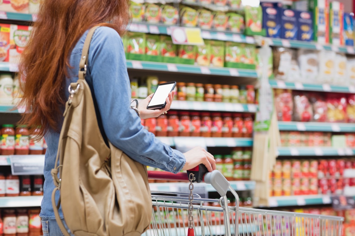 young woman with red hair holding a tan purse and cell phone and pushing a cart while grocery shopping