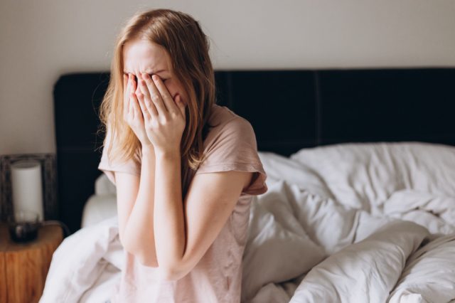 Woman sits on the bed and holds her head in her hands.