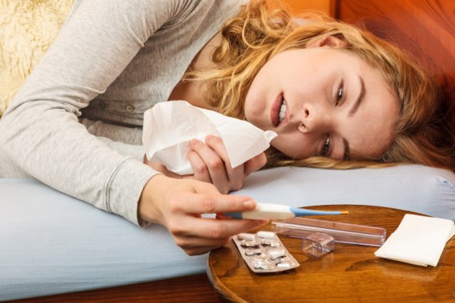 Sick woman laying in bed under wool blanket holding thermometer and tissue. Ill girl caught cold flu. Pills and tablets on table.