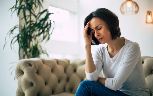 young woman, who is sitting on a sofa with her eyes closed, touching her head while suffering from a migraine.