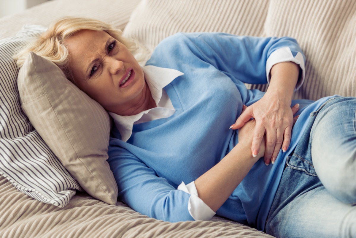 Mature woman in casual clothes is having a stomach ache while lying on couch at home.