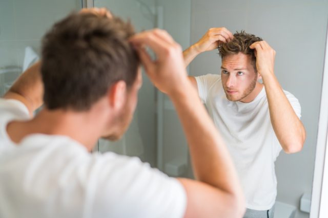 Young man in white t-shirt and jeans looking at thinning hair in mirror