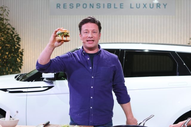 jamie oliver in blue shirt standing in front of a white car holding up a burger in one hand