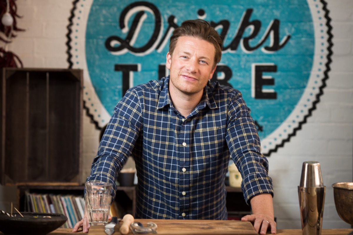 jamie oliver in blue plaid shirt standing in front of blue circular wooden sign reading "drinks"