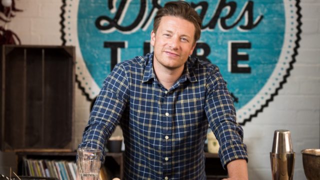 jamie oliver in blue plaid shirt standing in front of blue circular wooden sign reading "drinks"