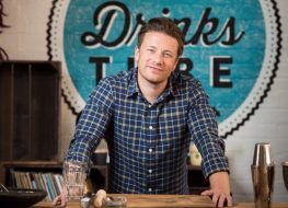 jamie oliver in blue plaid shirt standing in front of blue circular wooden sign reading "drinks"
