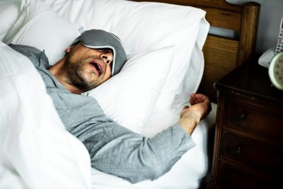 middle aged man in gray shirt and sleep mask sleeping with mouth open