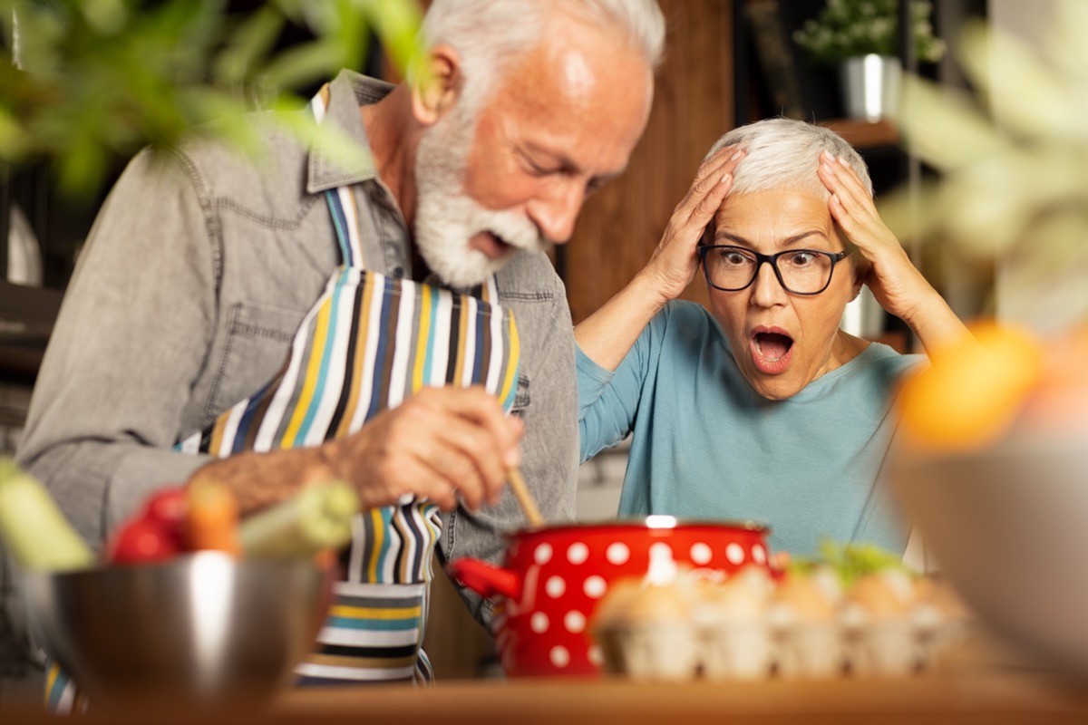 Senior woman shocked, with open mouth looking at her husband cooking.
