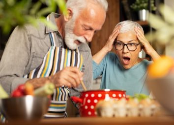 Senior woman shocked, with open mouth looking at her husband cooking.