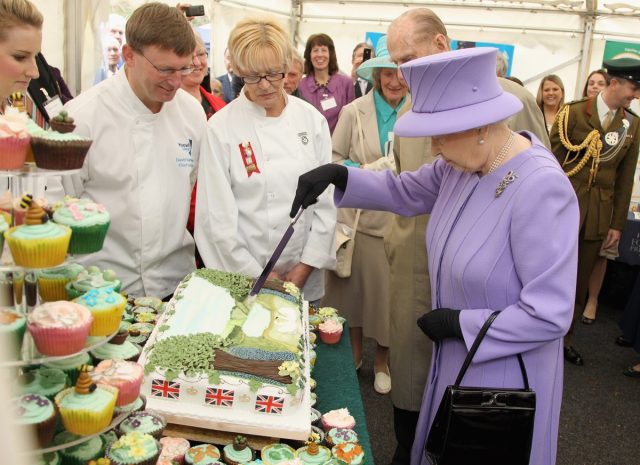 queen elizabeth in a purple suit cutting cake