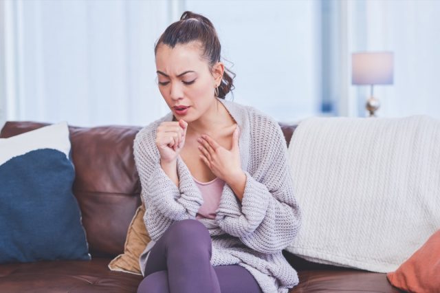 Young woman sitting alone on her couch at home and coughing.