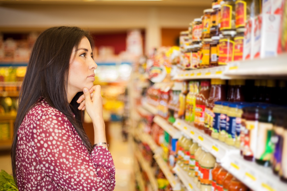 young woman looking at shelf of sauces appearing confused at grocery store