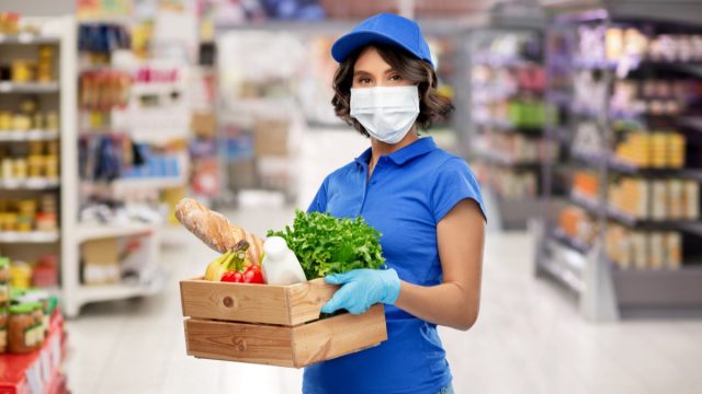 Delivery woman in face protective medical mask and gloves holding wooden box with food in supermarke.