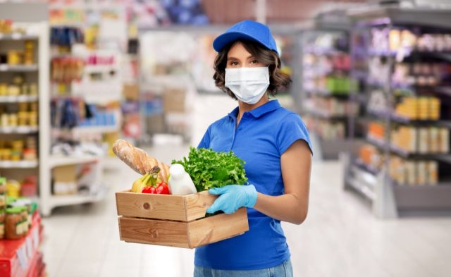 Delivery woman in face protective medical mask and gloves holding wooden box with food in supermarke.