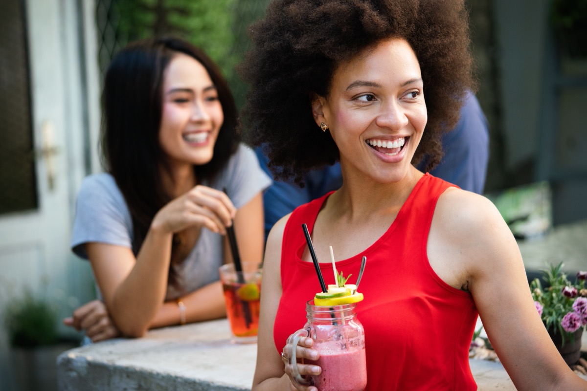 young woman outdoors in red shirt drinking smoothie while her friend behind her drinks iced tea