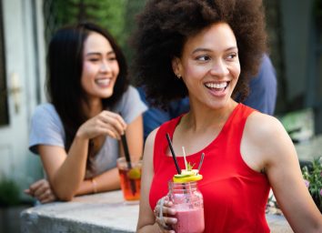 young woman outdoors in red shirt drinking smoothie while her friend behind her drinks iced tea