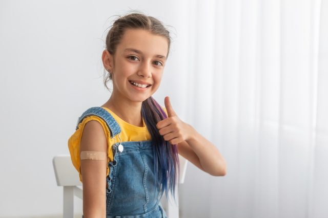 Happy smiling teenage patient showing vaccinated arm with patch on shoulder after getting shot and thumbs up gesture.