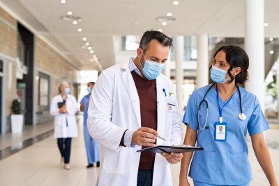 General practitioner and young nurse wearing surgical face mask against covid-19 while having a discussion in hospital hallway. Doctor with face mask discussing patient case status with his medical staff while walking on corridor. Worried busy doctor showing medical report to nurse and wearing protective face mask with copy space.