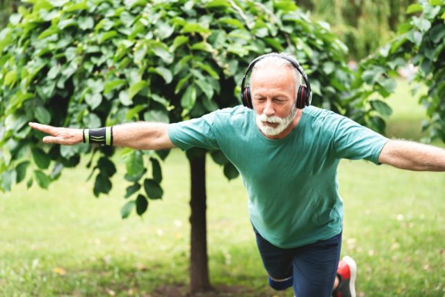 older man standing on one leg outdoors with headphones on
