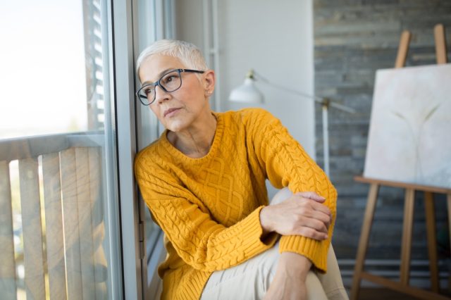 Sad mature woman sitting on the floor of her living room and looking through the window.