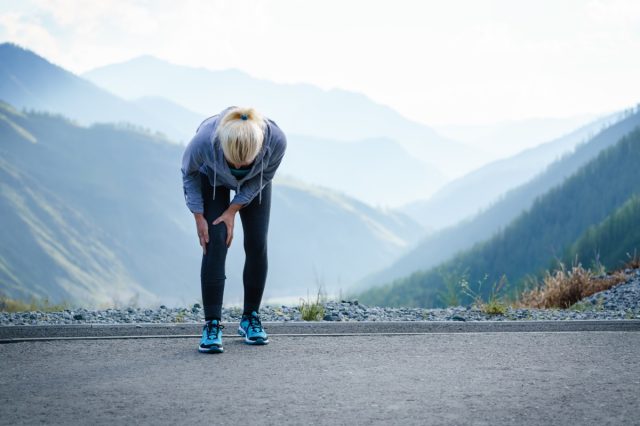 mujer con dolor de rodilla caminando por un sendero con montañas detrás de ella