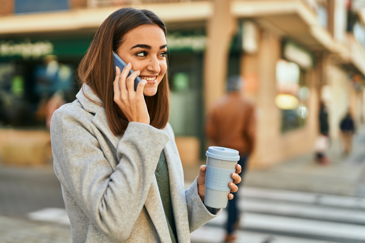 young woman drinking coffee outdoors while on cell phone