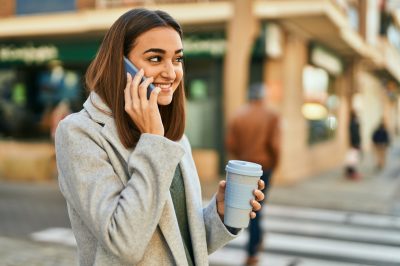 young woman drinking coffee outdoors while on cell phone