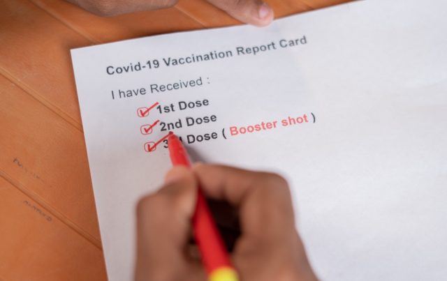 Close up of hands checking Covid-19 vaccine card and ticking 3rd or booster dose after vaccination.