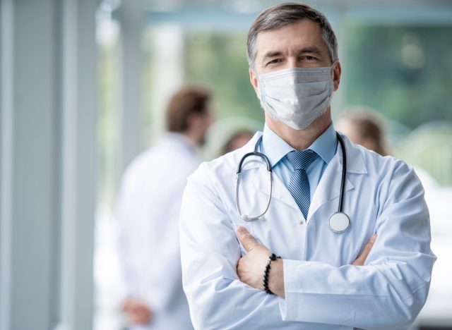 Cheerful mature doctor in face mask posing at camera, healthcare and medicine.