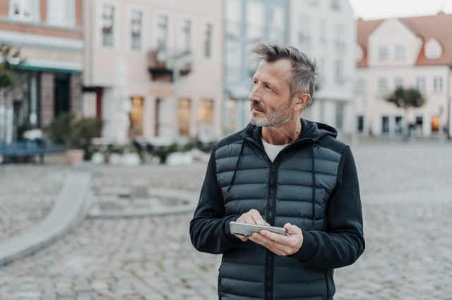 Troubled middle-aged man staring off the his right as he stands in a deserted square in a historic town holding his mobile phone in his hand.