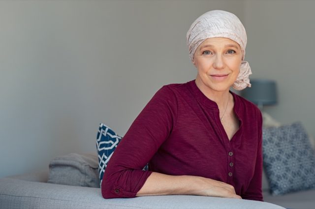 Reife Frau mit Krebs in rosa Kopfschmuck lächelnd auf dem Sofa zu Hause sitzen