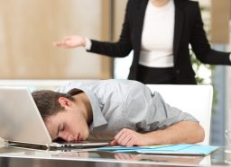 Employee sleeping with the face over the laptop with his angry boss watching back at office