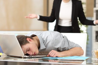Employee sleeping with the face over the laptop with his angry boss watching back at office