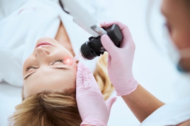 Female doctor in latex gloves performing the laser skin resurfacing on the young patient face