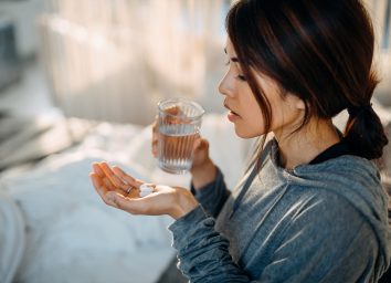 Young Asian woman sitting on bed and feeling sick, taking medicines in hand with a glass of water.