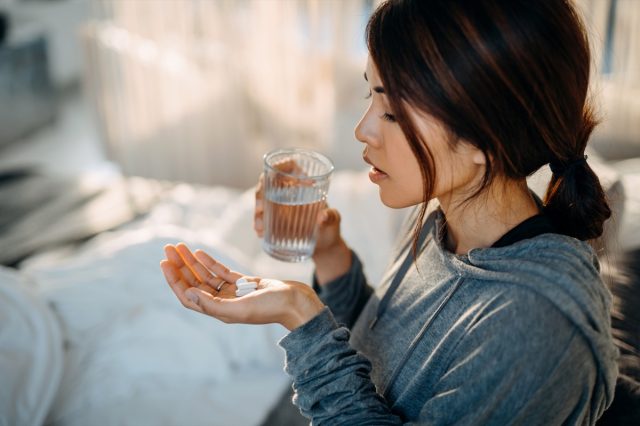 Young Asian woman sitting on bed and feeling sick, taking medicines in hand with a glass of water.