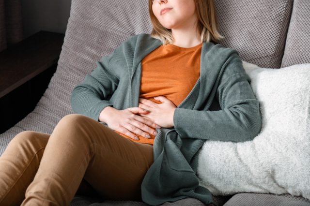Young woman suffers, writhes in abdominal pain lying on the couch in the living room at home interior
